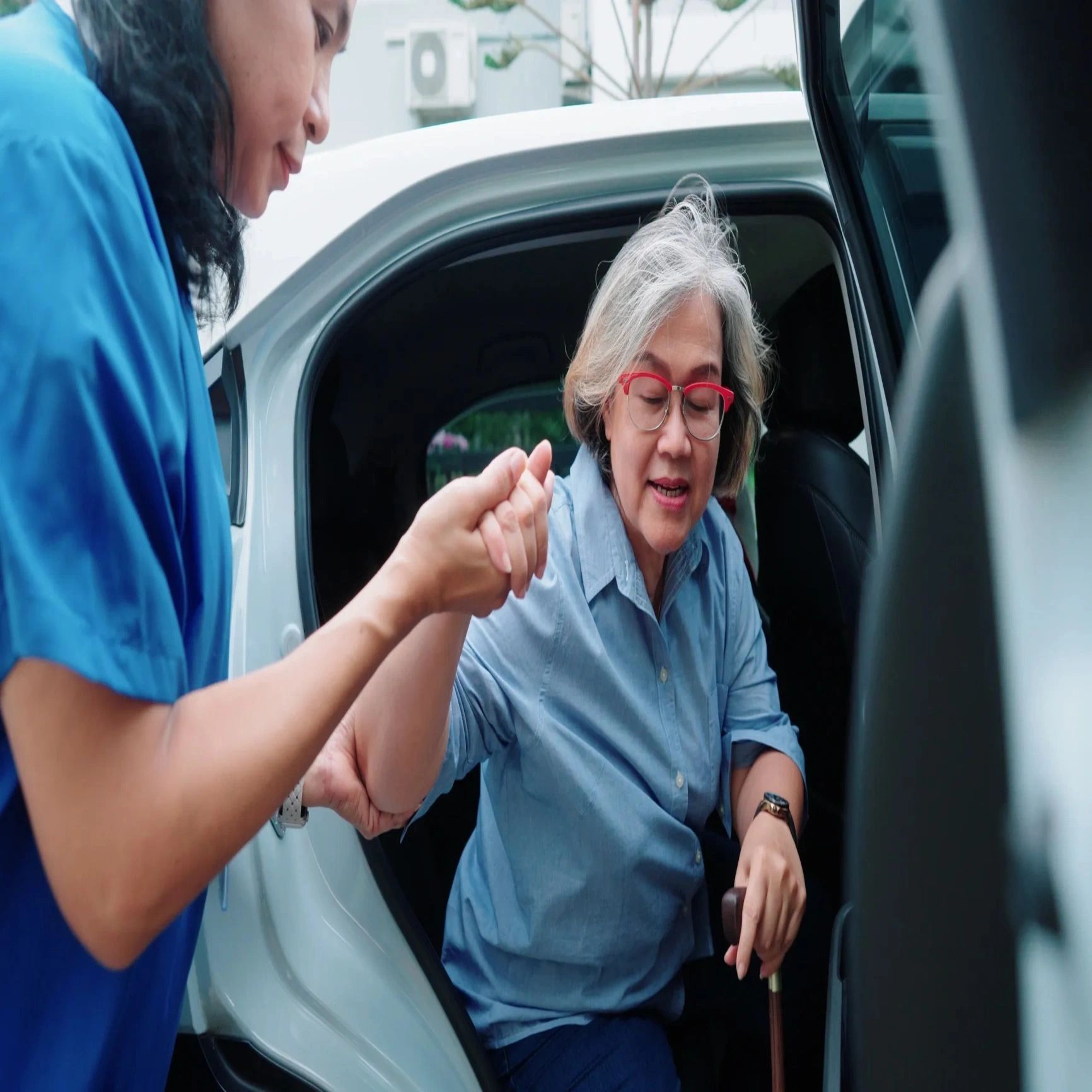 Caregiver helping elderly woman with glasses get out of a car.
