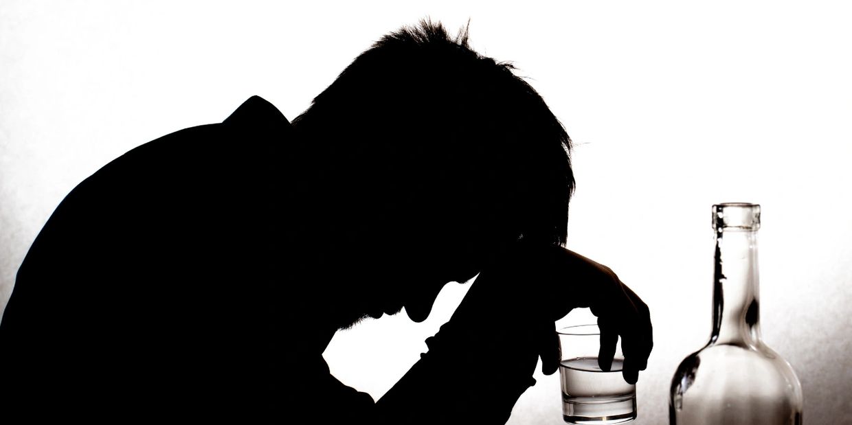 A man sits alone at a table, holding a glass of alcohol. He is contemplating recovery from addiction