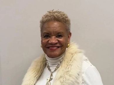Smiling woman wearing a white fur vest and jewelry.