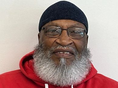 Smiling elderly man with a full gray beard and glasses wearing a red hoodie and black beanie.