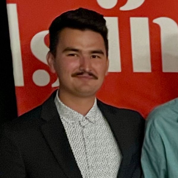 Man in a black suit and patterned white shirt smiling in front of a red backdrop.