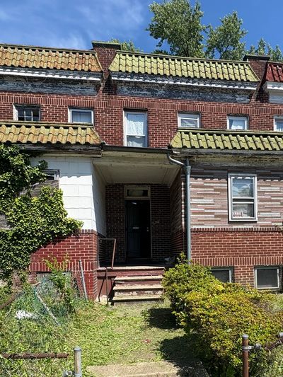 A brick row house with a tiled roof and an overgrown front yard under a clear blue sky.