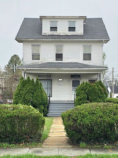 Old white house with a gray roof and green bushes in front.