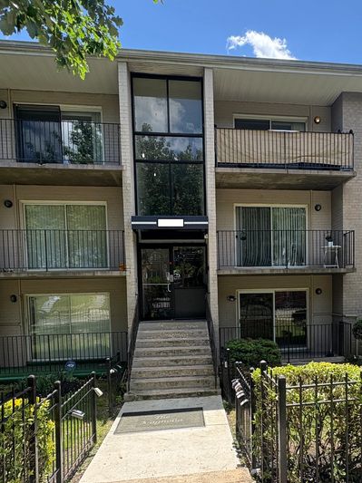 Entrance of a three-story residential apartment building with balconies and greenery.