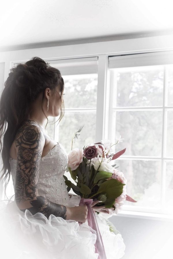 Bride with tattoos holding a bouquet near a window.