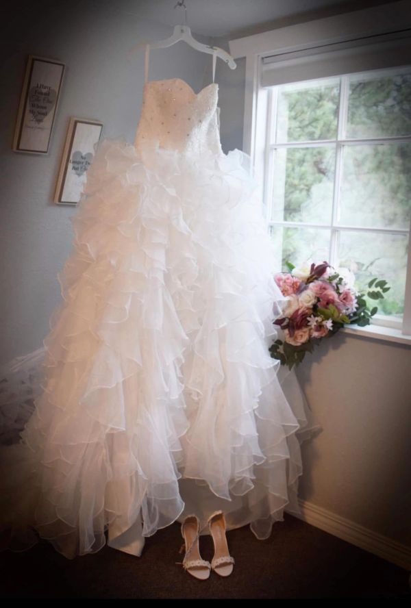 Elegant layered white wedding dress hanging near window with bouquet.