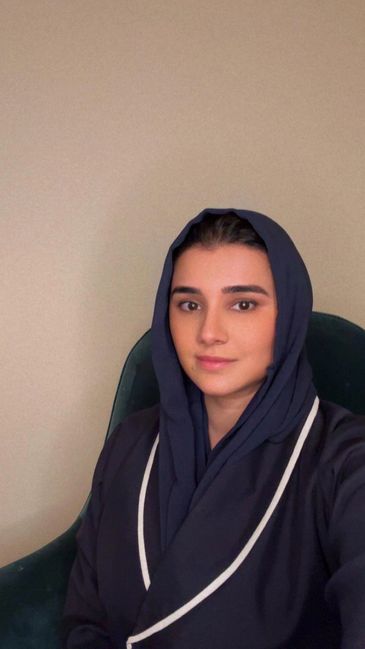 A woman wearing a navy blue hijab and matching outfit, seated indoors.