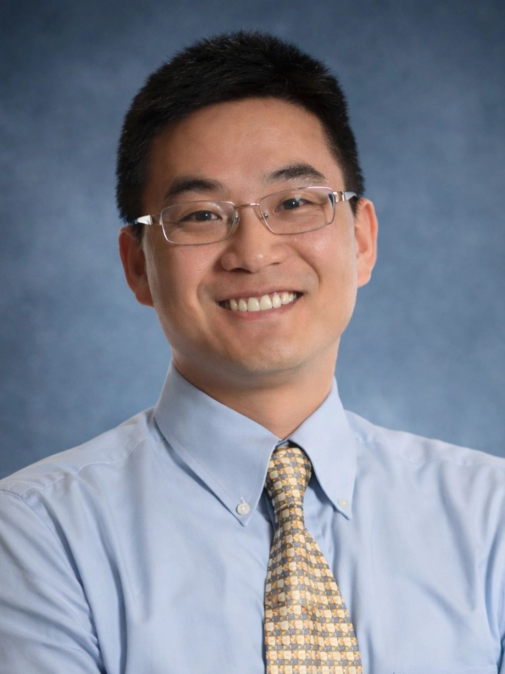 Man in glasses smiling in a blue shirt and patterned tie.