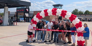 Group celebrates ribbon-cutting event under a red and white balloon arch.