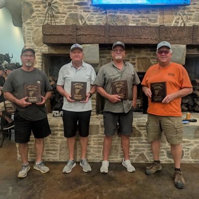 Four men holding award plaques in front of a stone fireplace.