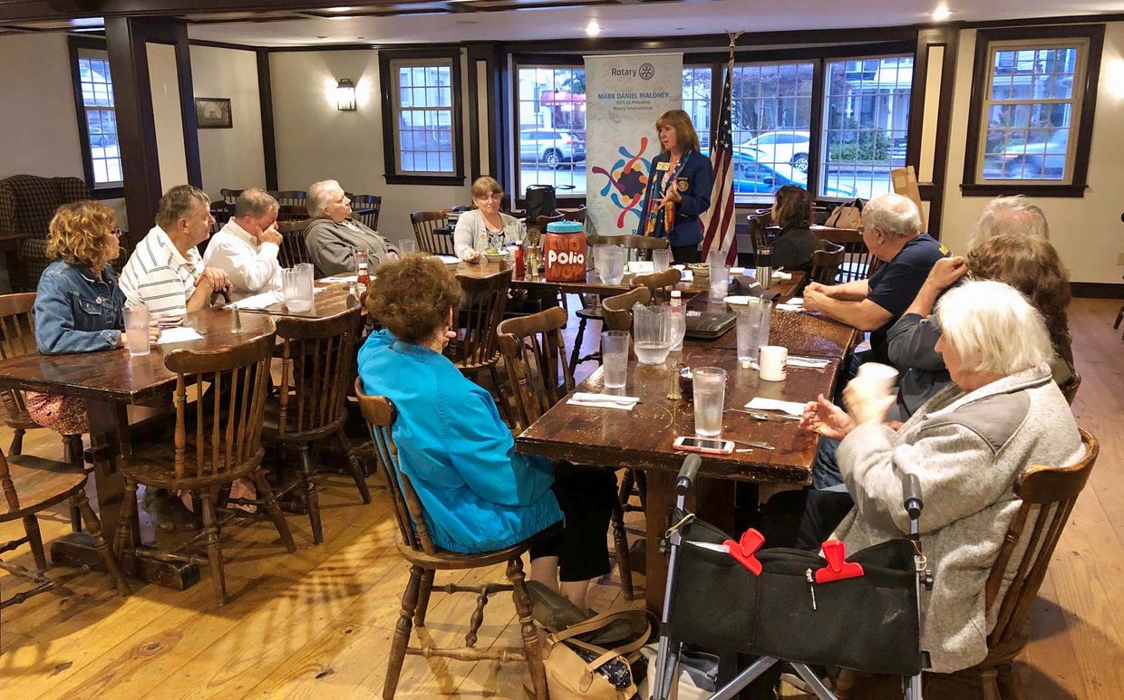 A woman speaks to a group at a Rotary Club meeting in a cozy room.