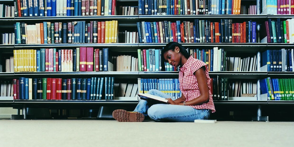 Young woman reading a book while sitting on the library floor.