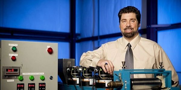 Man in a shirt and tie posing with industrial machinery and control panel.