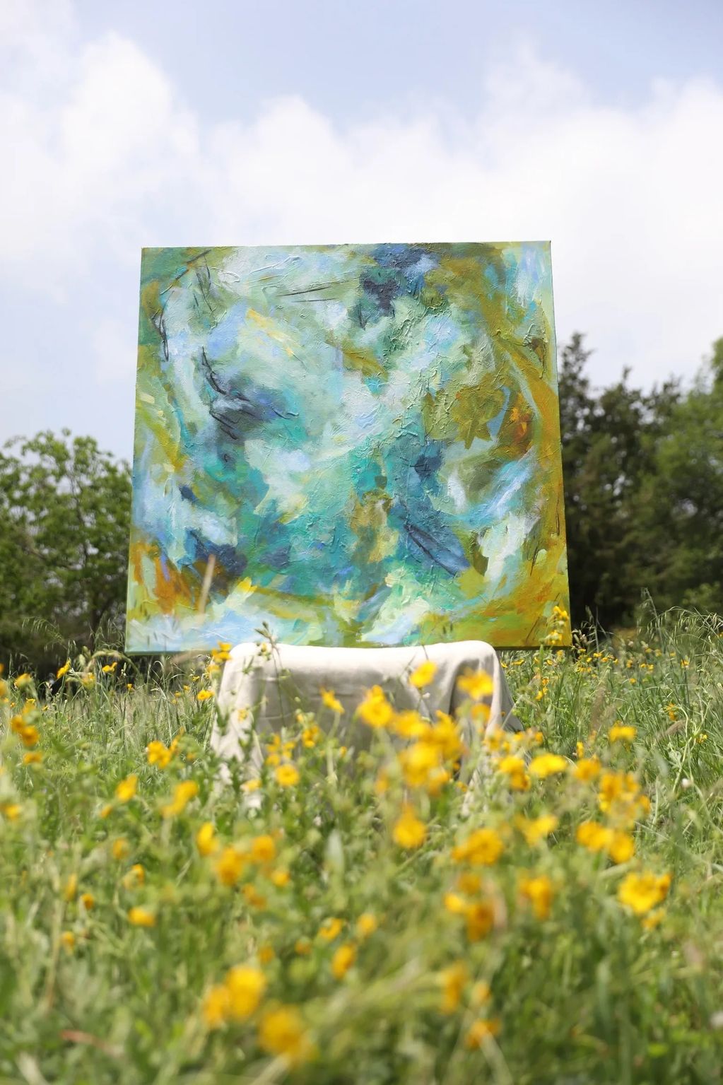 Abstract painting displayed outdoors among yellow wildflowers under a partly cloudy sky.