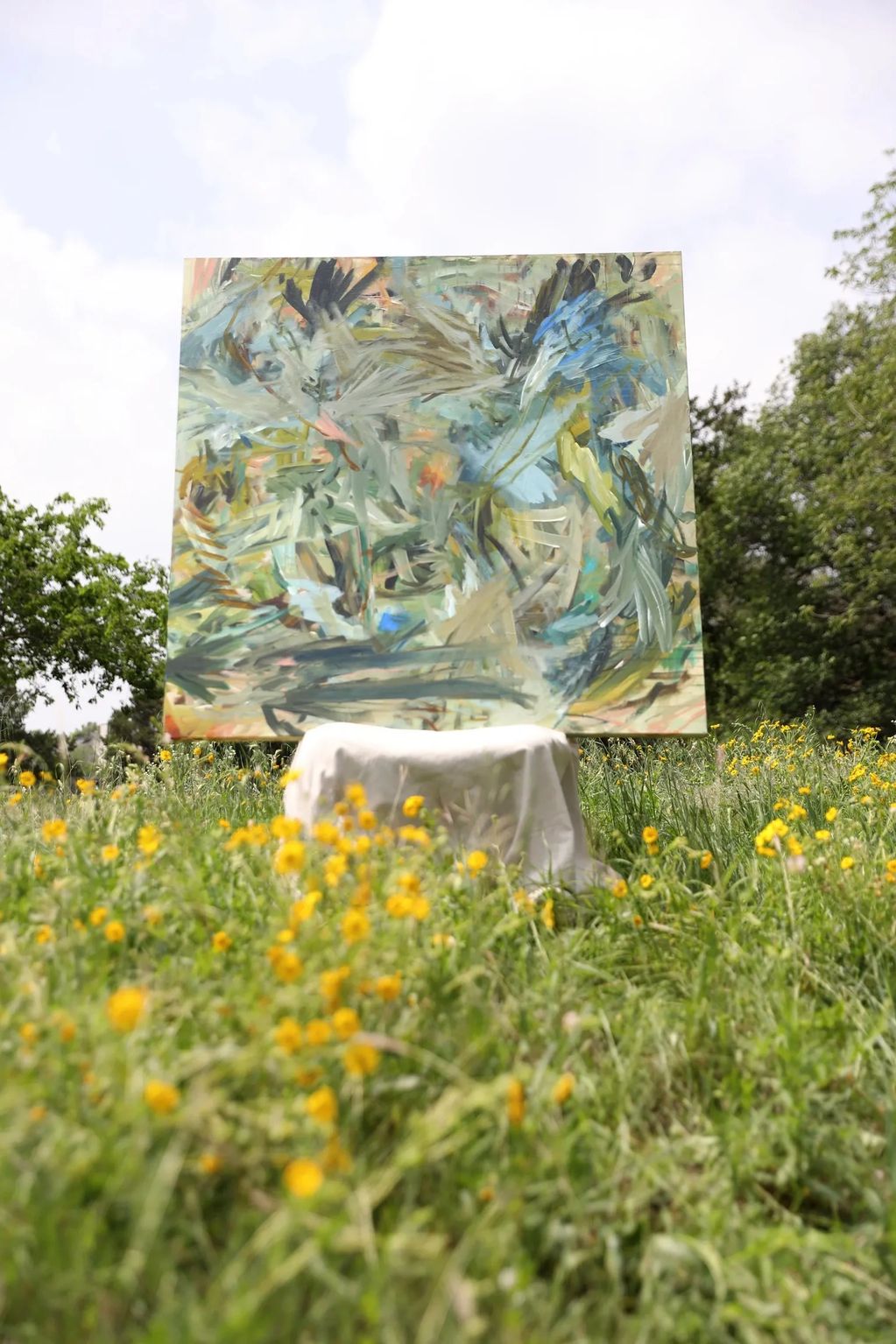 Abstract painting displayed outdoors amidst yellow wildflowers and greenery.