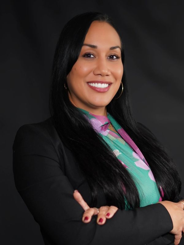 Confident woman in a black blazer smiling with arms crossed against a dark background.