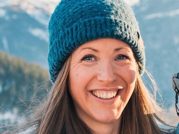 Smiling woman in a blue knit hat with snowy mountains in the background.