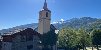 Church tower and stone house with snowy mountains in background under clear blue sky. in Seez 