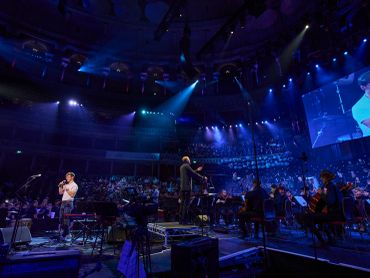 A live orchestral concert with a conductor and a large audience under dramatic lighting.