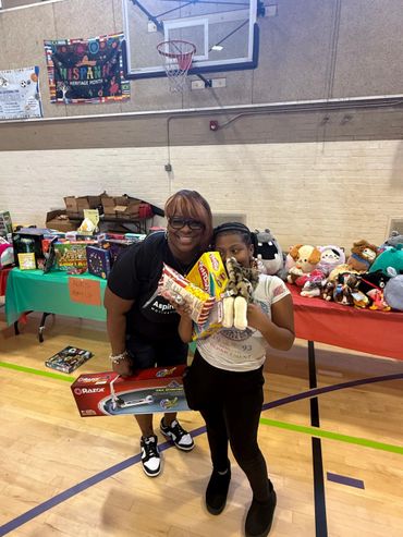 Woman and child holding toys and snacks in a gym with donation tables.