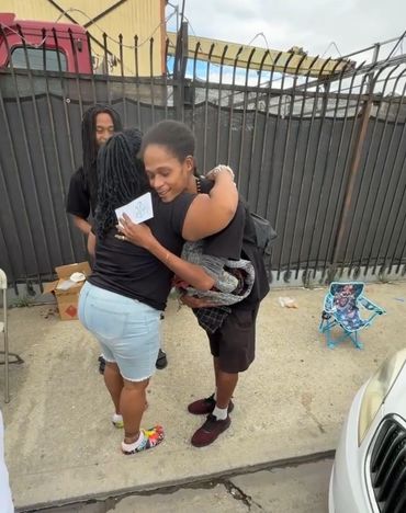 Two women warmly hugging outdoors near a fence, with a man standing nearby.