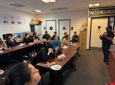 A woman giving a presentation to a group in a classroom with a 'NO EXCUSES' sign.