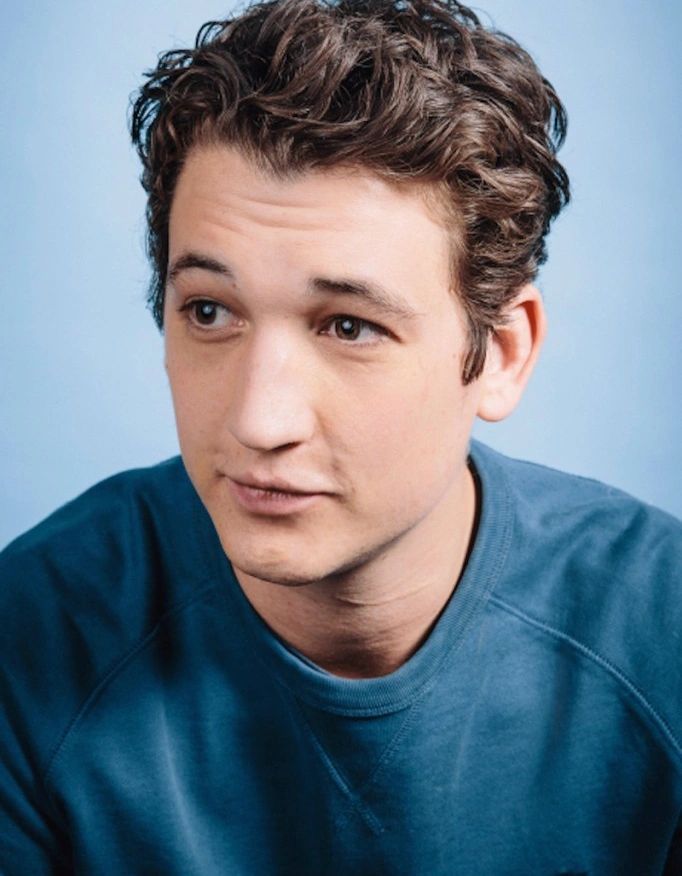 A young man with curly hair wearing a blue sweater looks thoughtfully to the side.