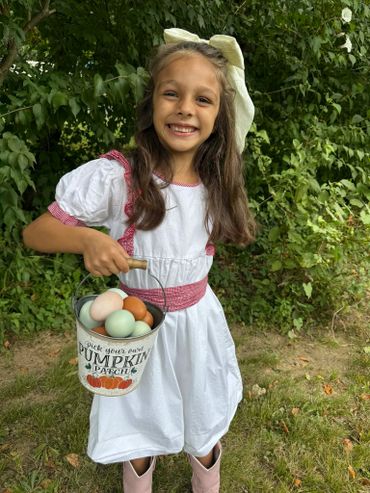 Young girl smiling, holding a bucket of colorful eggs in a pumpkin patch.