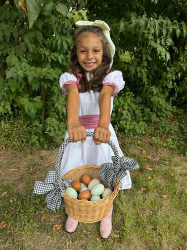 Smiling girl in white dress holding a basket of colorful eggs outdoors.