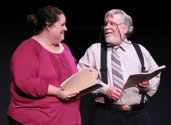 Two actors rehearsing lines together on stage, holding scripts.