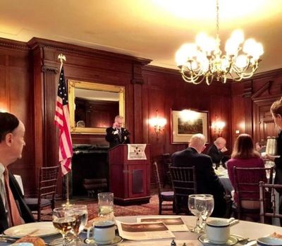 Formal event in a wood-paneled room with an American flag and speaker at podium.