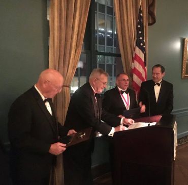 Four men in tuxedos at a formal event with an American flag in the background.