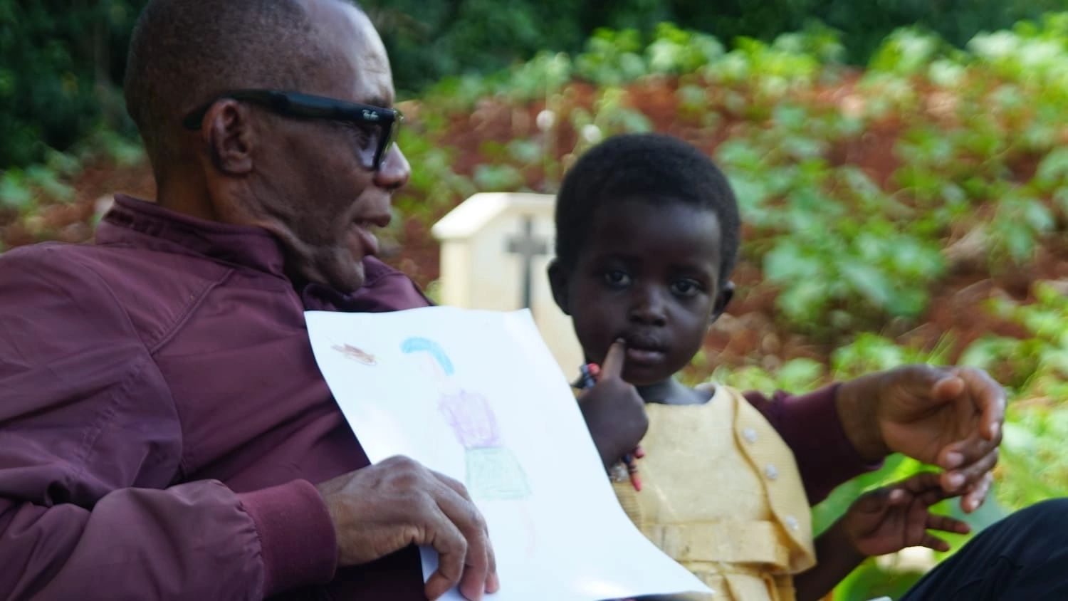 Man shows drawing to a young child outdoors.