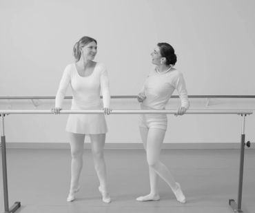 Two female ballet dancers at the barre, smiling and chatting in a dance studio.