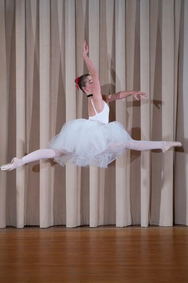 A ballerina in a white tutu leaps gracefully in front of beige curtains.