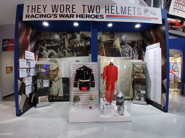 Exhibit honoring racing drivers who served in the military, featuring helmets and uniforms.