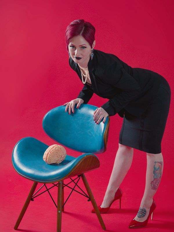 Woman in black with red hair leans on blue chair with a brain model.