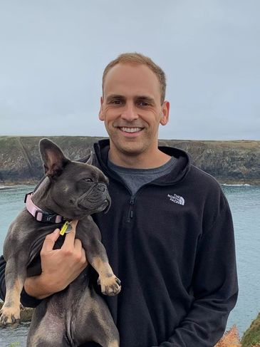 Man holding a black French bulldog by the water on a cloudy day.