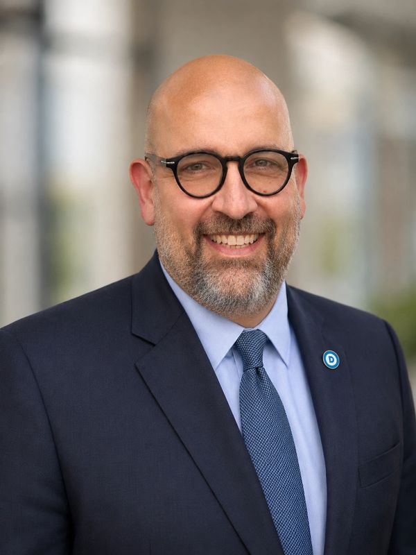 Smiling man in glasses and suit with a blue tie and Democratic pin.
