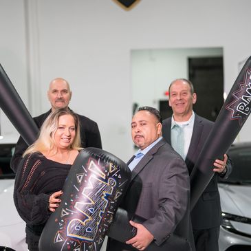 THREE MEN AND A LADY HOLDING INFLATABLE HAMMERS THAT READ "BAM!" IN FRONT OF TWO LUXURY SPORT CARS.