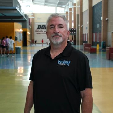 Man in a black Venom Athletics shirt standing in a spacious hallway.