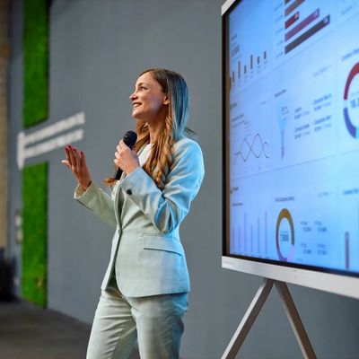 Woman in suit giving a presentation with charts on a screen.
