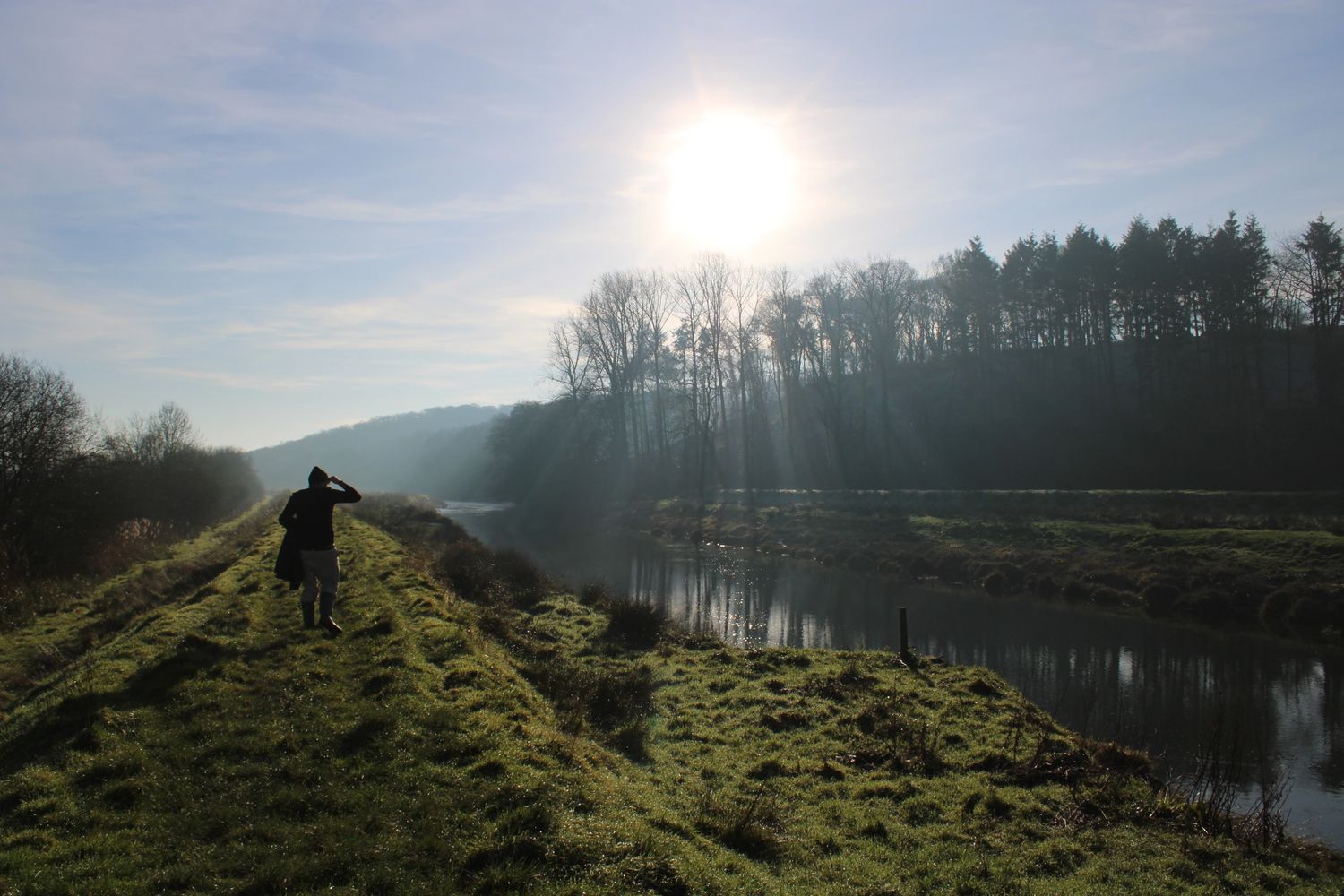 Person walking along a grassy riverbank under a bright sun.