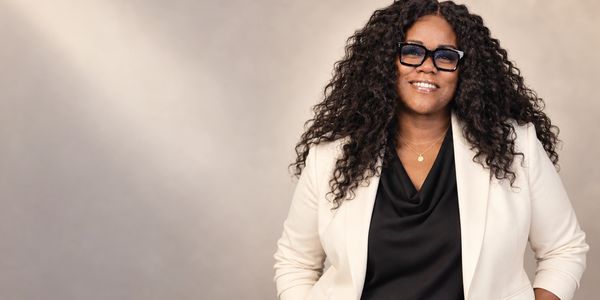 Smiling woman with curly hair wearing glasses and a white blazer.