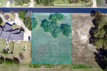A vacant grassy lot with some trees, bordered by a paved road and neighboring houses.