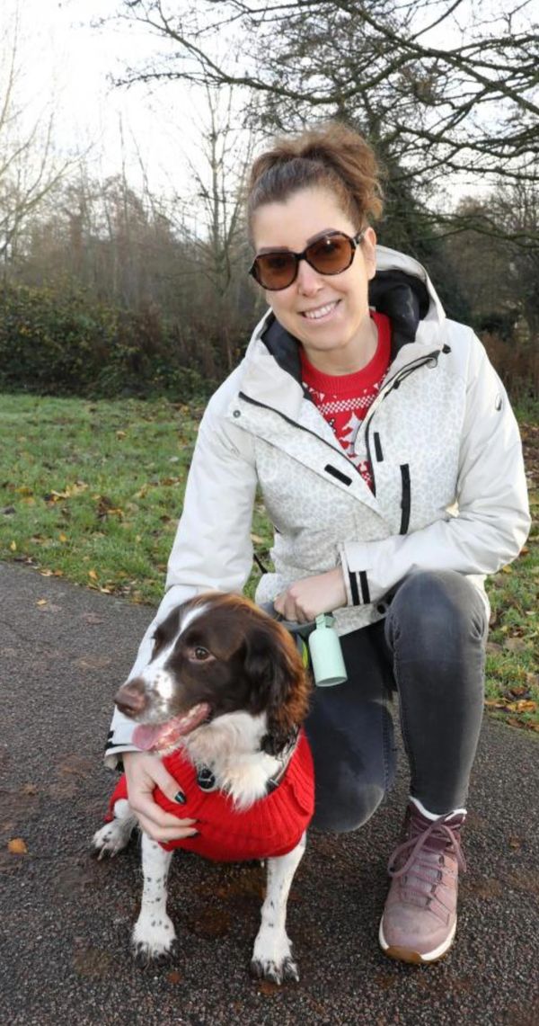 Woman in sunglasses kneeling with her dog wearing a red sweater outdoors.