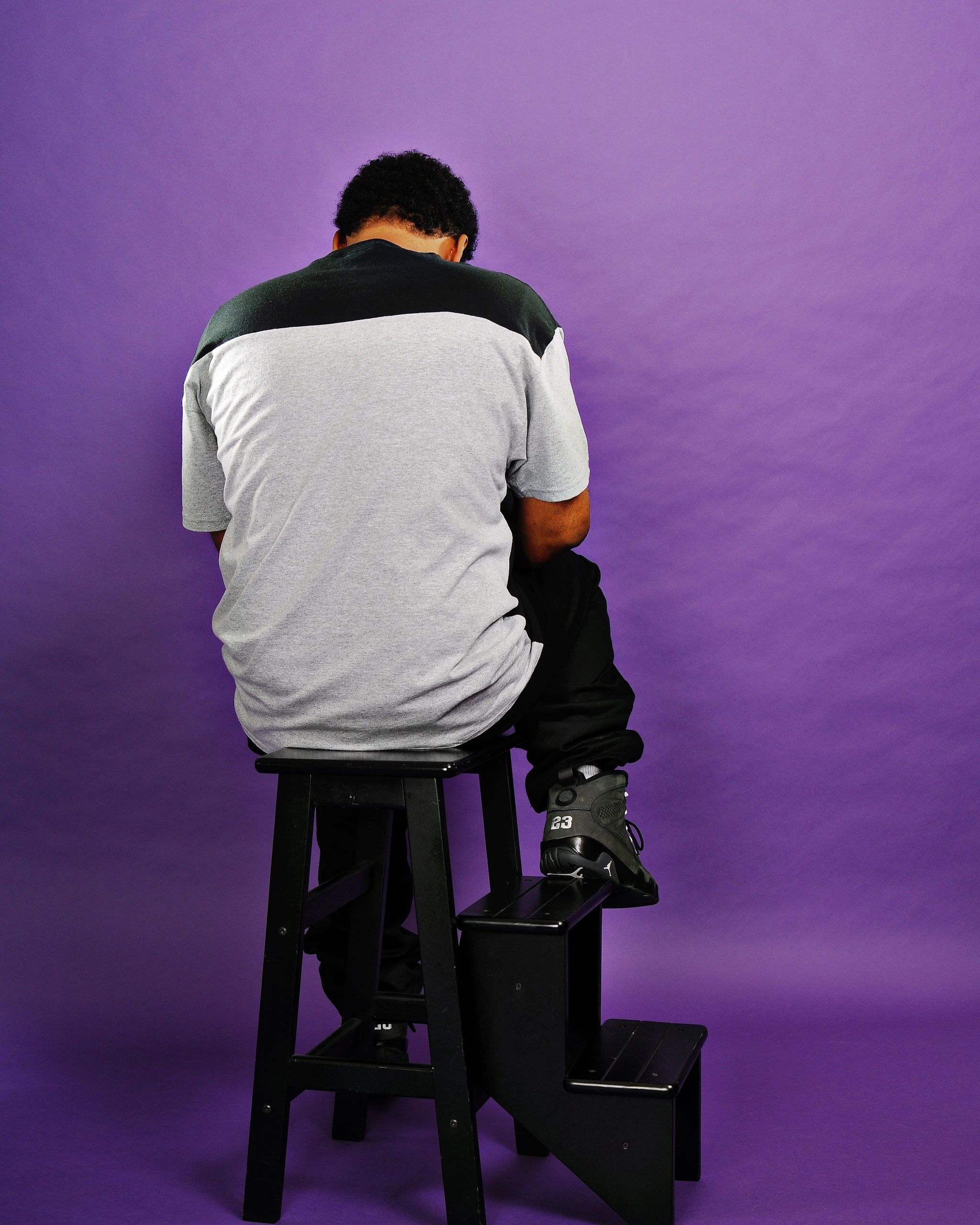Person sitting on a black stool against a purple backdrop, facing away.