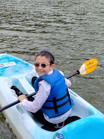 A smiling boy wearing sunglasses and a blue life jacket paddles a kayak on a lake.
