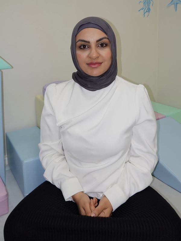 Woman wearing a grey hijab and white blouse sitting calmly indoors.