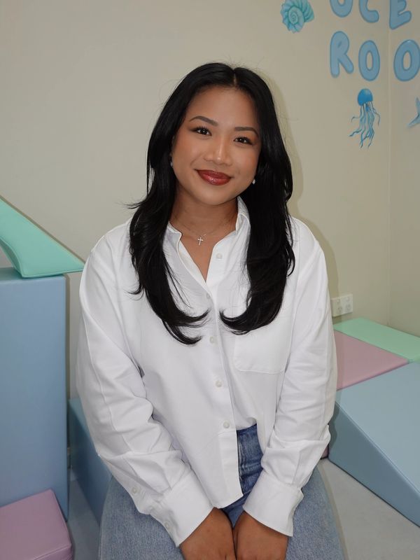 A smiling woman in a white shirt and jeans sits in a pastel-themed room.